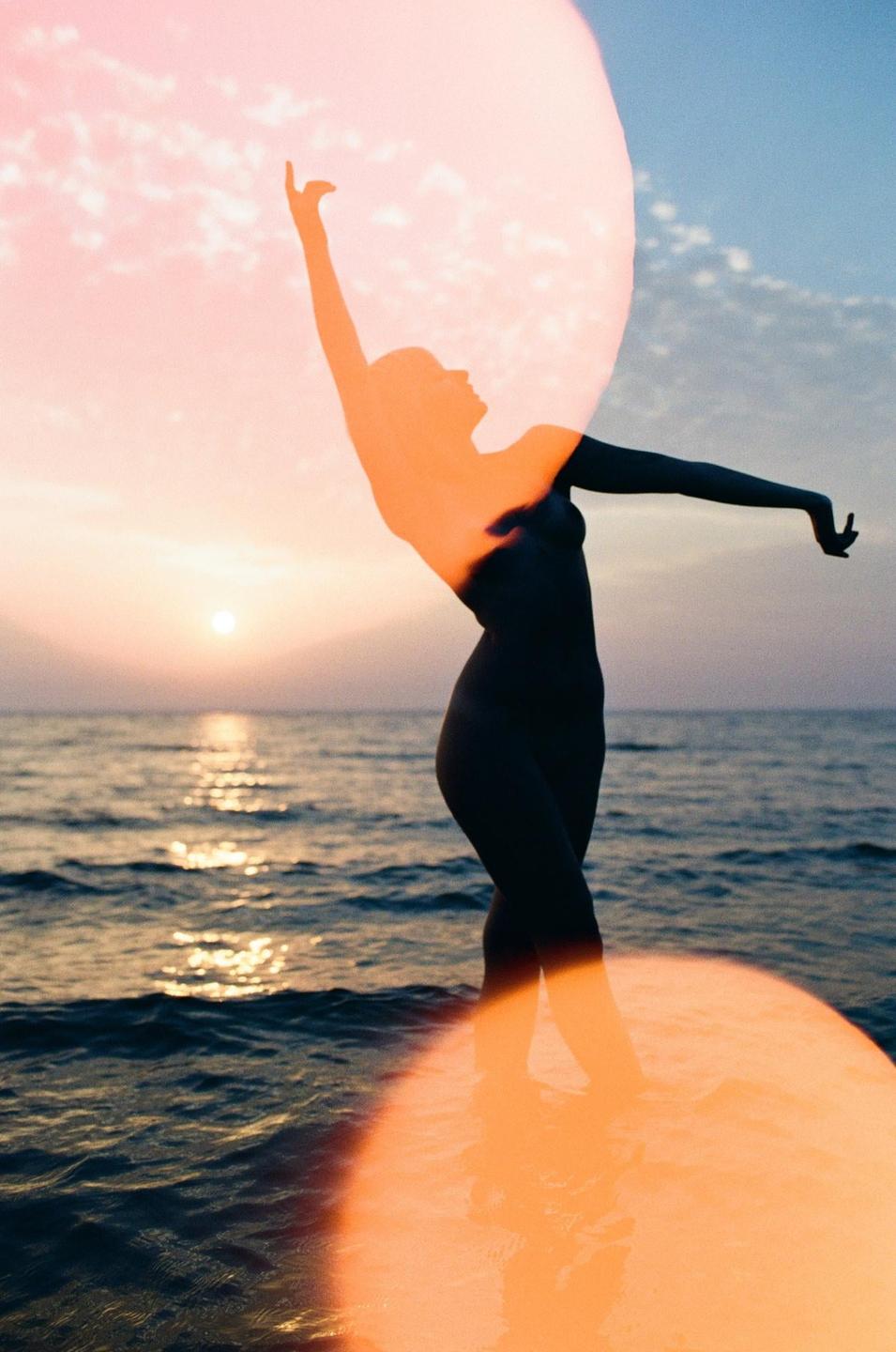 A woman stands in the ocean with her arms raised, silhouetted against a sunset sky with a large orange light leak.