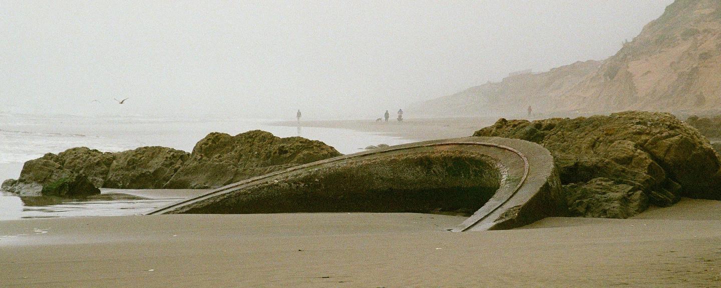A concrete structure resembling a sunken ship is partially buried in the sand on a foggy beach with people and rocks in the distance.