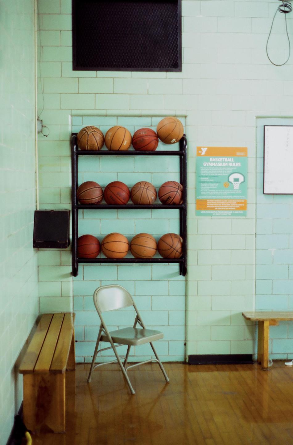 A rack of basketballs stands against a tiled wall in a gymnasium, with a folding chair and wooden benches nearby.