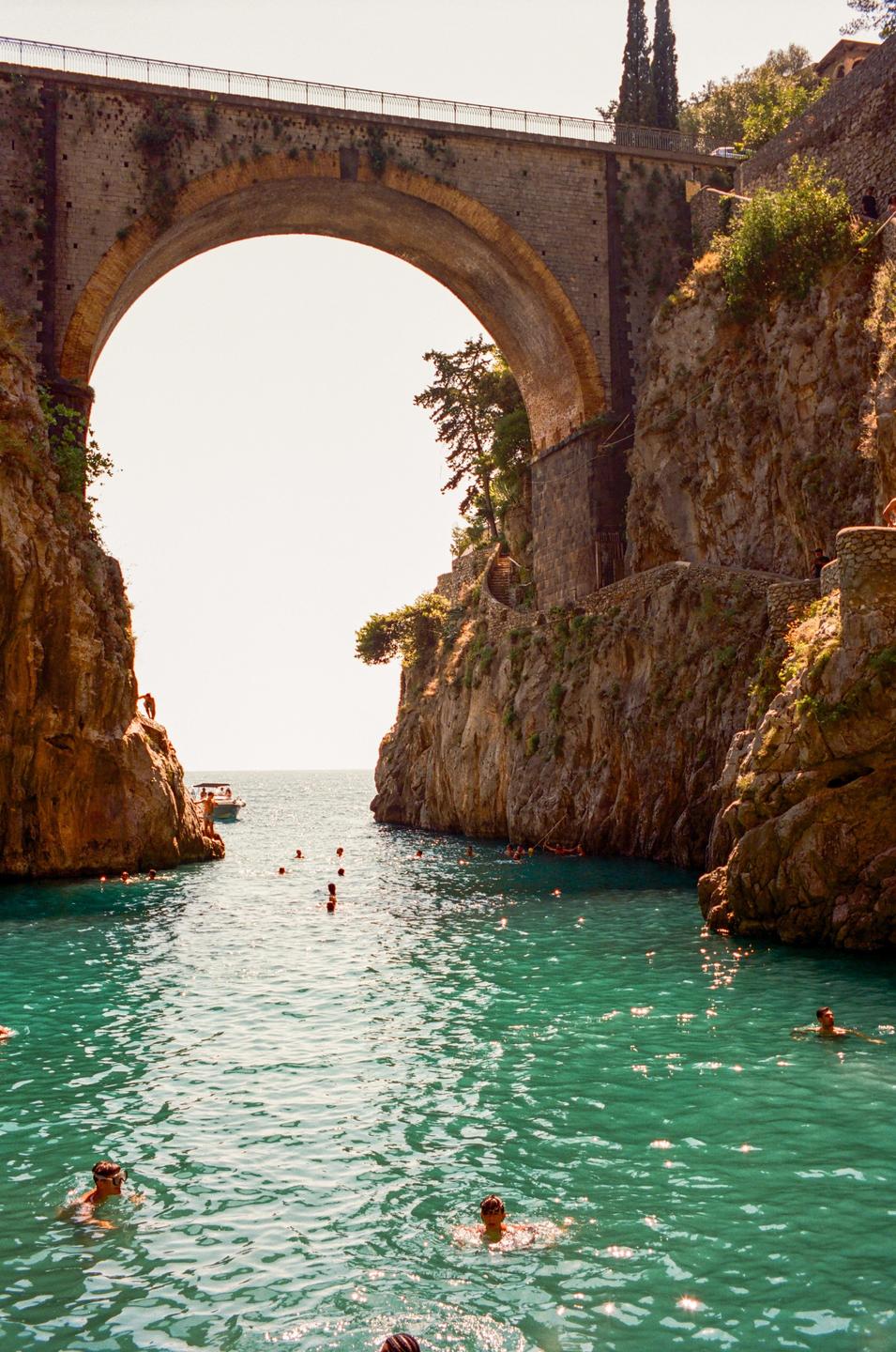 A stone bridge arches over a narrow inlet where people are swimming in turquoise water.