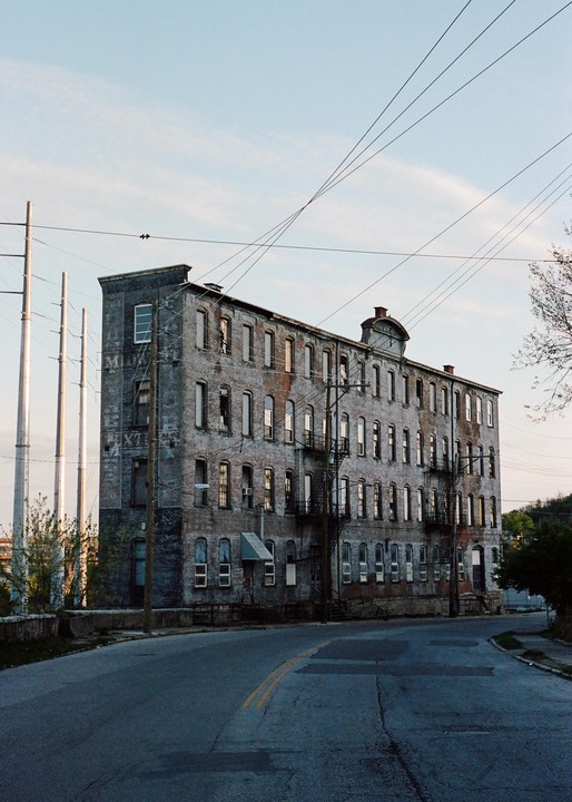 An old brick building with many windows stands next to a road with power lines overhead.