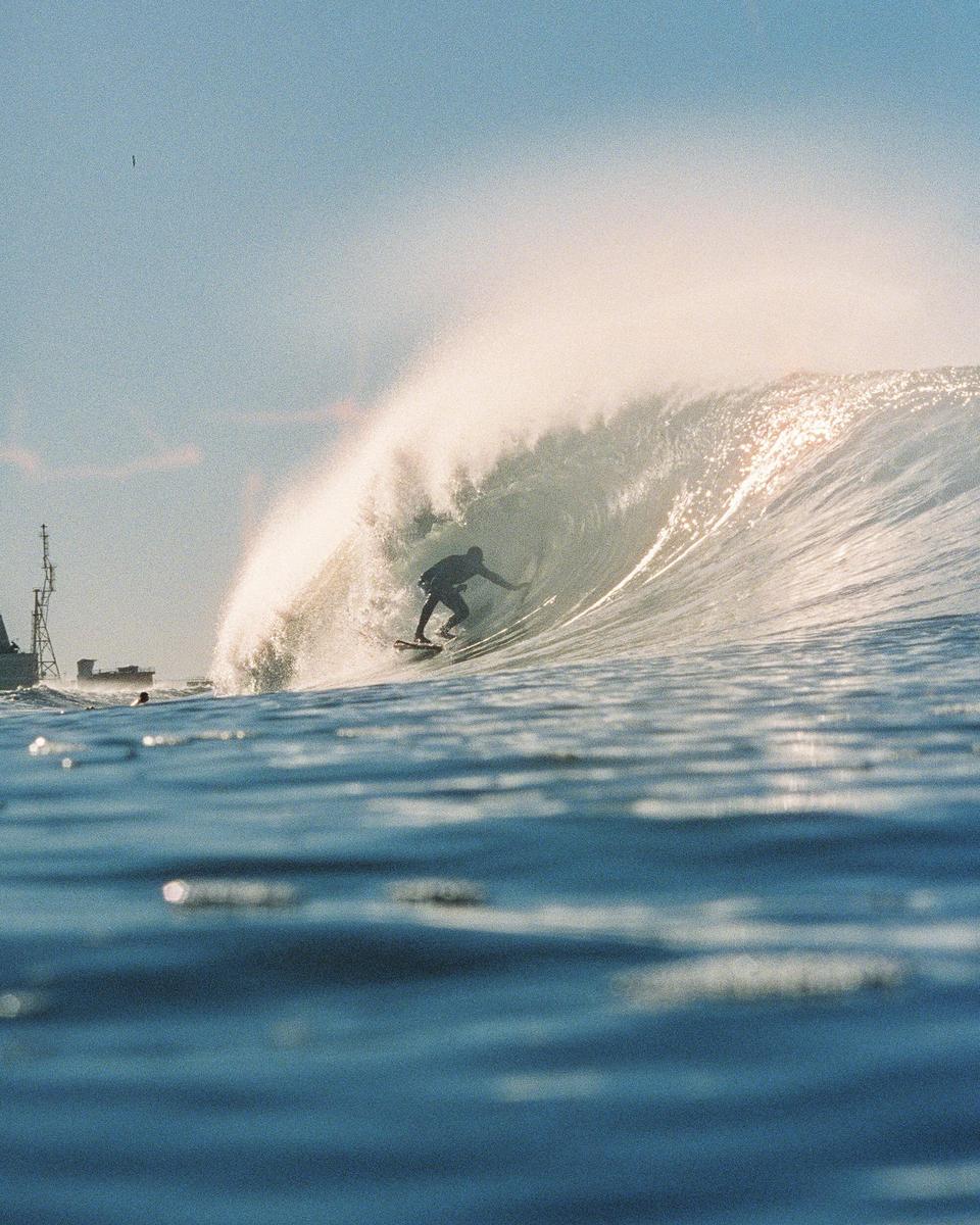 A surfer rides a large wave with a ship visible in the background.