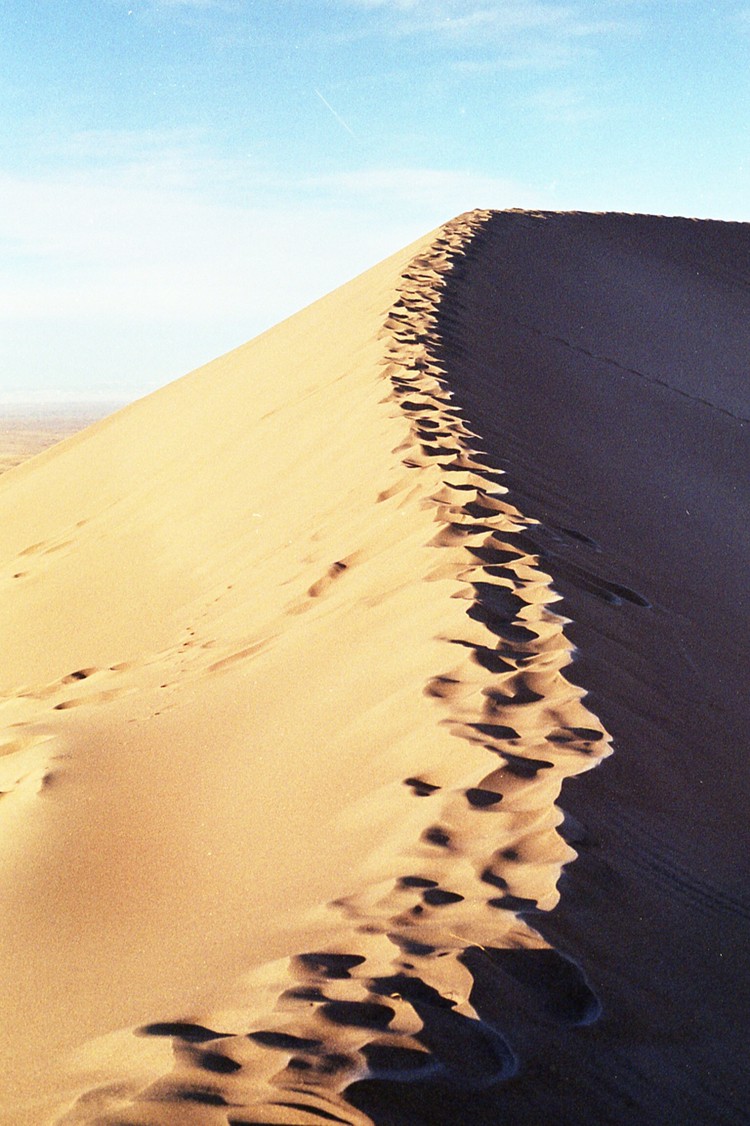 Footprints lead up the ridge of a sand dune under a clear sky.