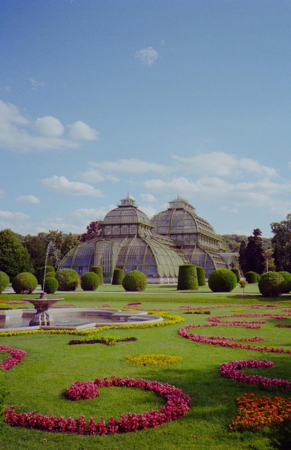 A large glass greenhouse stands behind a fountain and a manicured lawn with flowerbeds.