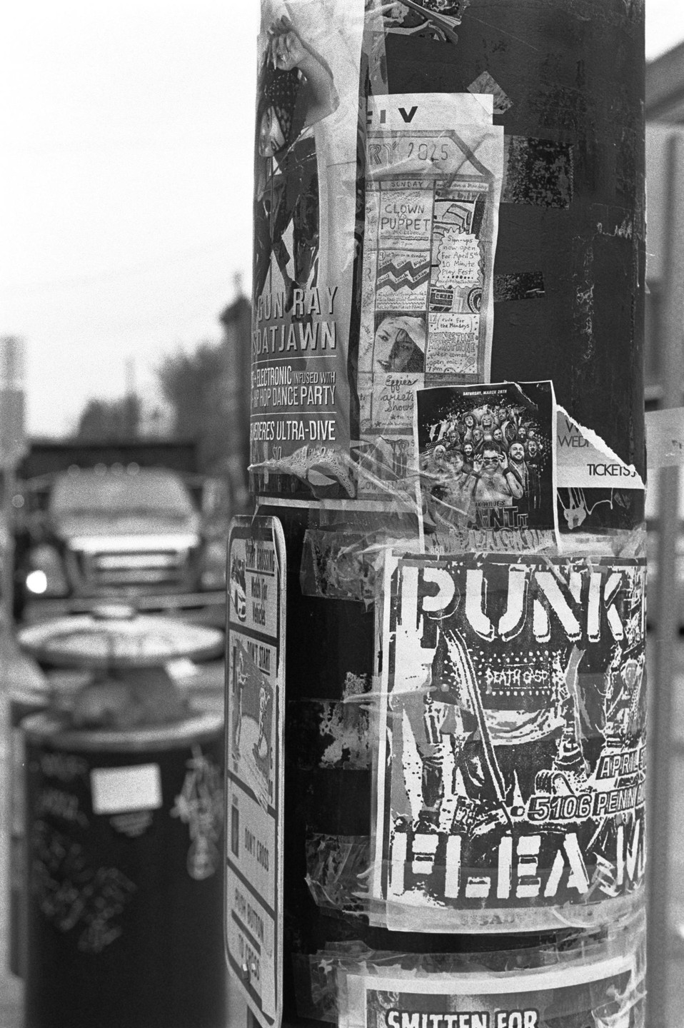 A street pole is covered in various posters and flyers, with a blurry street and vehicle in the background.