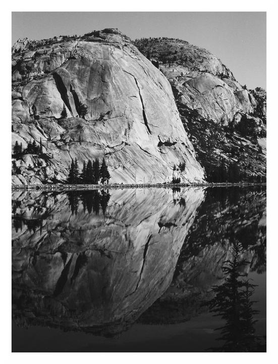 A large rock formation is reflected in the still water of a lake.