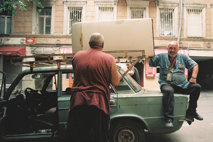 Two men are loading a refrigerator onto the roof rack of a green car while another man sits on the trunk.