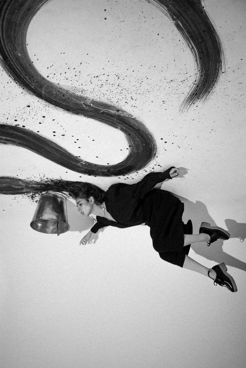 A woman in black clothing lies on a white surface with a bucket and a large black painted stroke.
