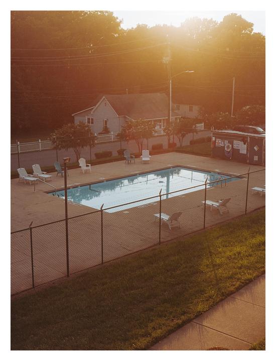 A fenced swimming pool with lounge chairs is visible from above, with houses and trees in the background under a bright sun.