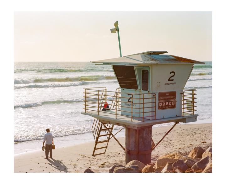 A man stands on a sandy beach next to a lifeguard tower as waves crash in the background.