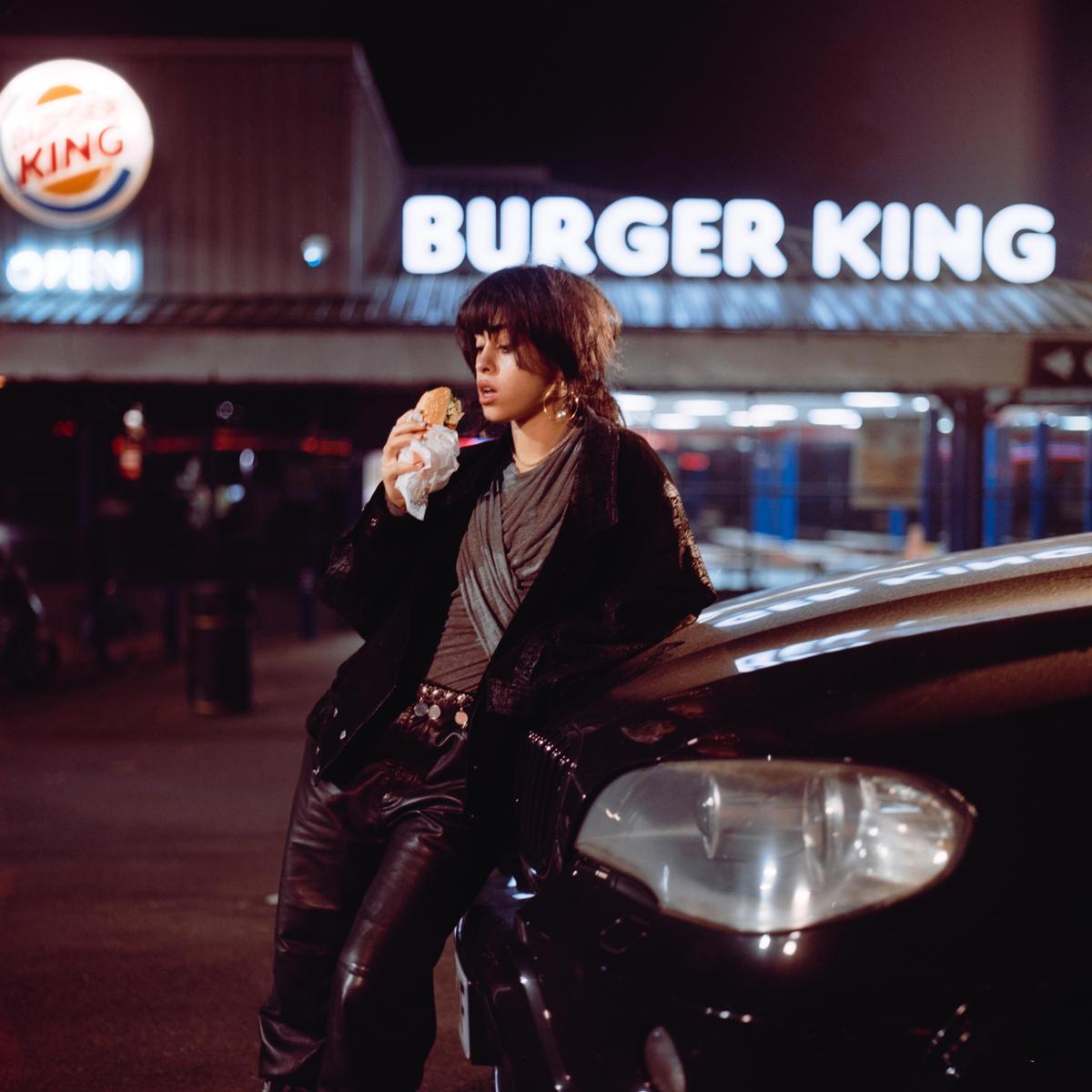 A woman eats a burger while leaning on a car at night with a Burger King restaurant in the background.