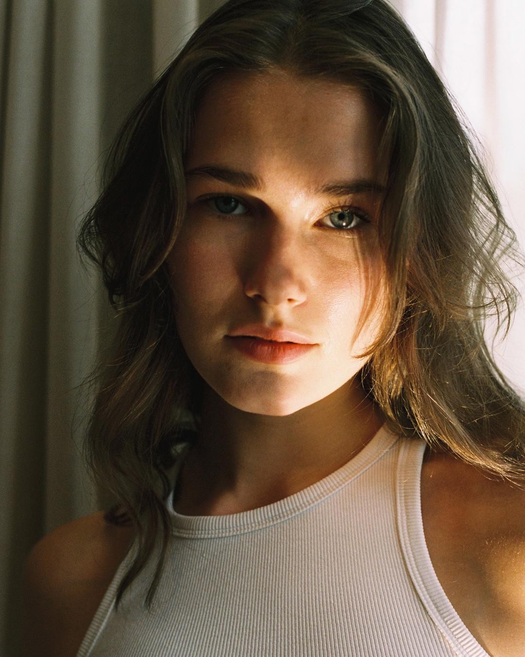 A woman with long hair and a white tank top looks directly at the viewer.
