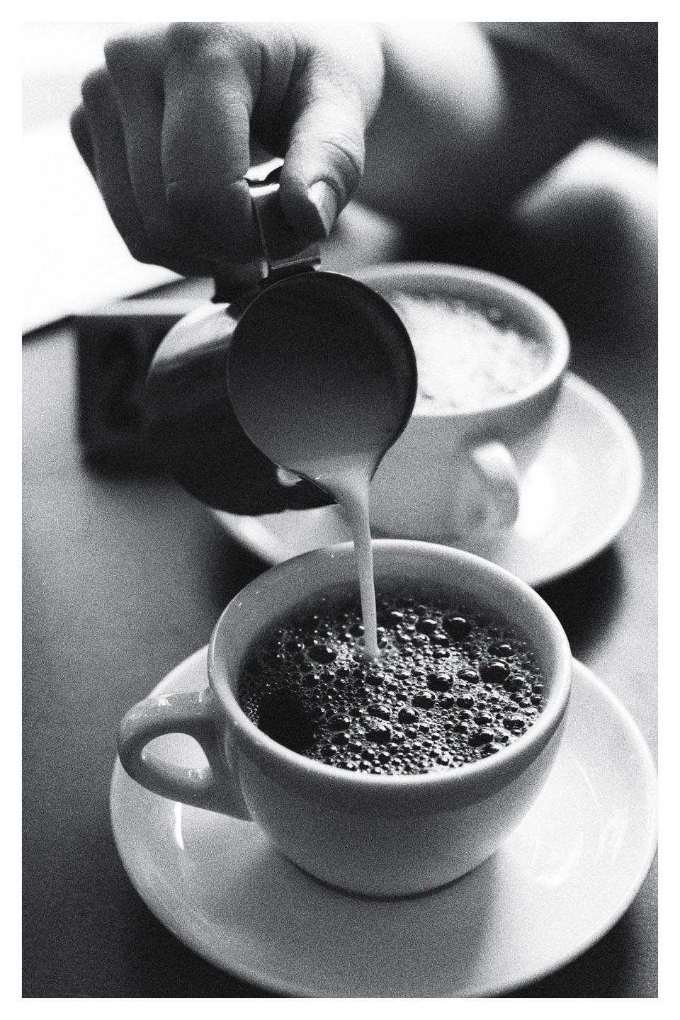 A hand pours milk into a cup of coffee, with another cup in the background.