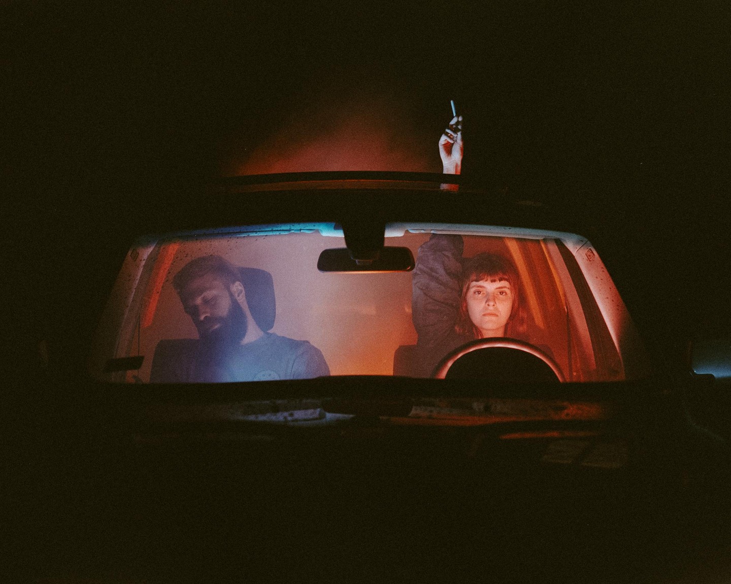 A woman looks forward from the driver's seat of a car at night while a man sleeps in the passenger seat and a hand holds a cigarette out of the sunroof.