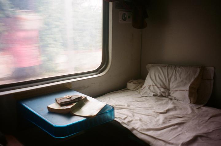 A bed and a small table with a book and wallet are next to a window on a moving train.