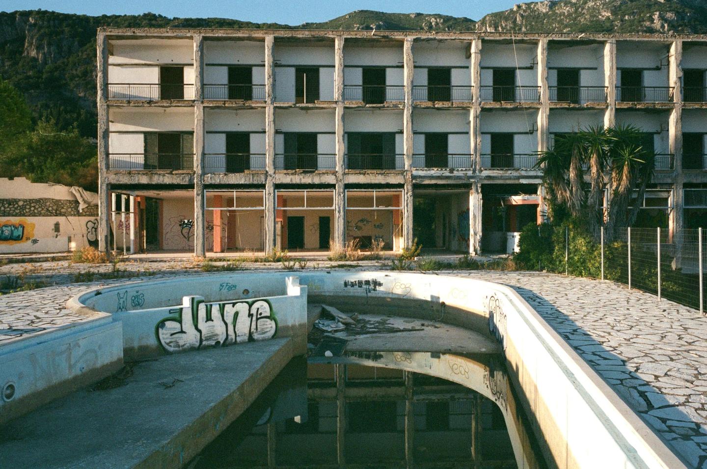 An abandoned hotel with a swimming pool in the foreground and mountains in the background is shown in a wide shot.