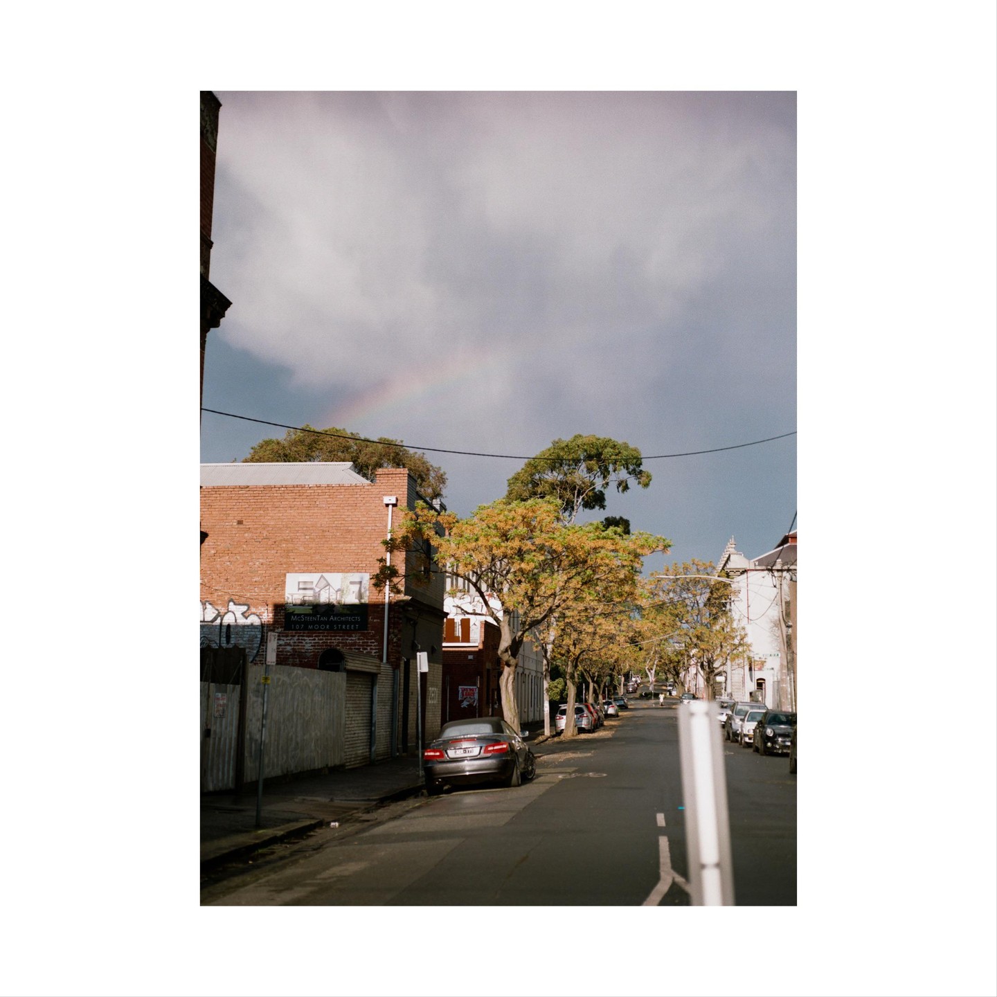A rainbow appears over a street lined with buildings and trees with a car parked on the left.