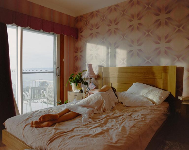 A woman lies in bed with her feet visible, next to a window overlooking a balcony and the sea.