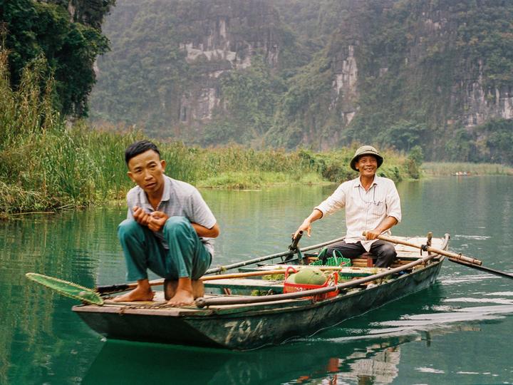 Two men are in a boat on a river with mountains and lush vegetation in the background.