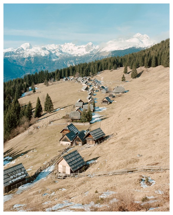 A village of wooden huts is nestled on a grassy hillside with snow-capped mountains in the background.