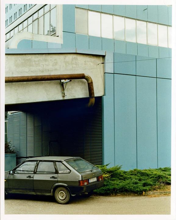 A gray car is parked next to a building with blue panels and a large rusty pipe.