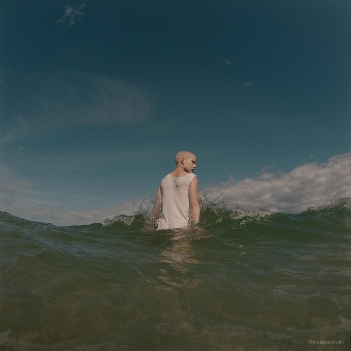 A person with a shaved head stands in the ocean as a wave breaks behind them.