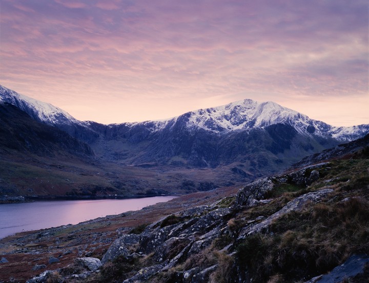 A snow-capped mountain range overlooks a lake under a pink and purple sky.