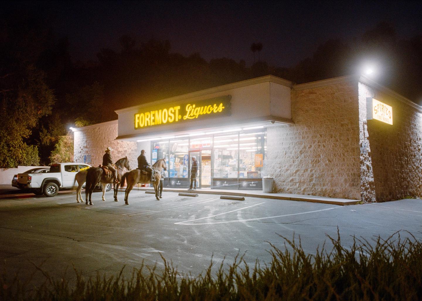 Three men on horseback are outside a liquor store at night, with a fourth man standing near the entrance.