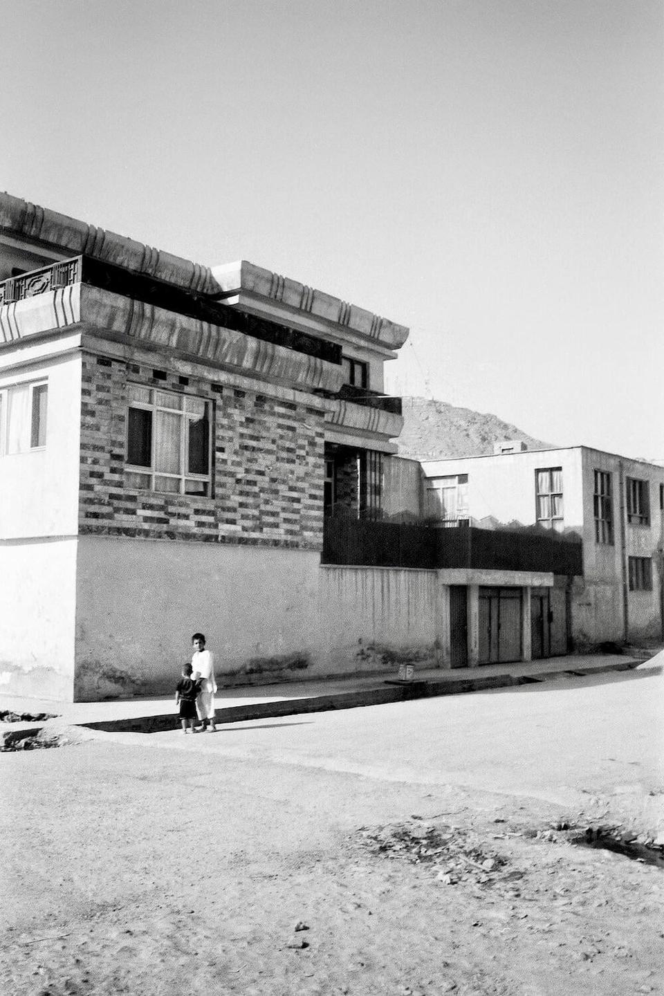 A woman and child stand on a sidewalk in front of a building with a mountain in the background.