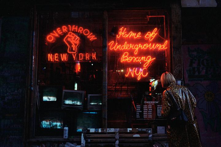A woman in a leopard print coat stands in front of a storefront with red neon signs that read "Overthrow New York" and "Home of Underground Boxing NYC" at night.