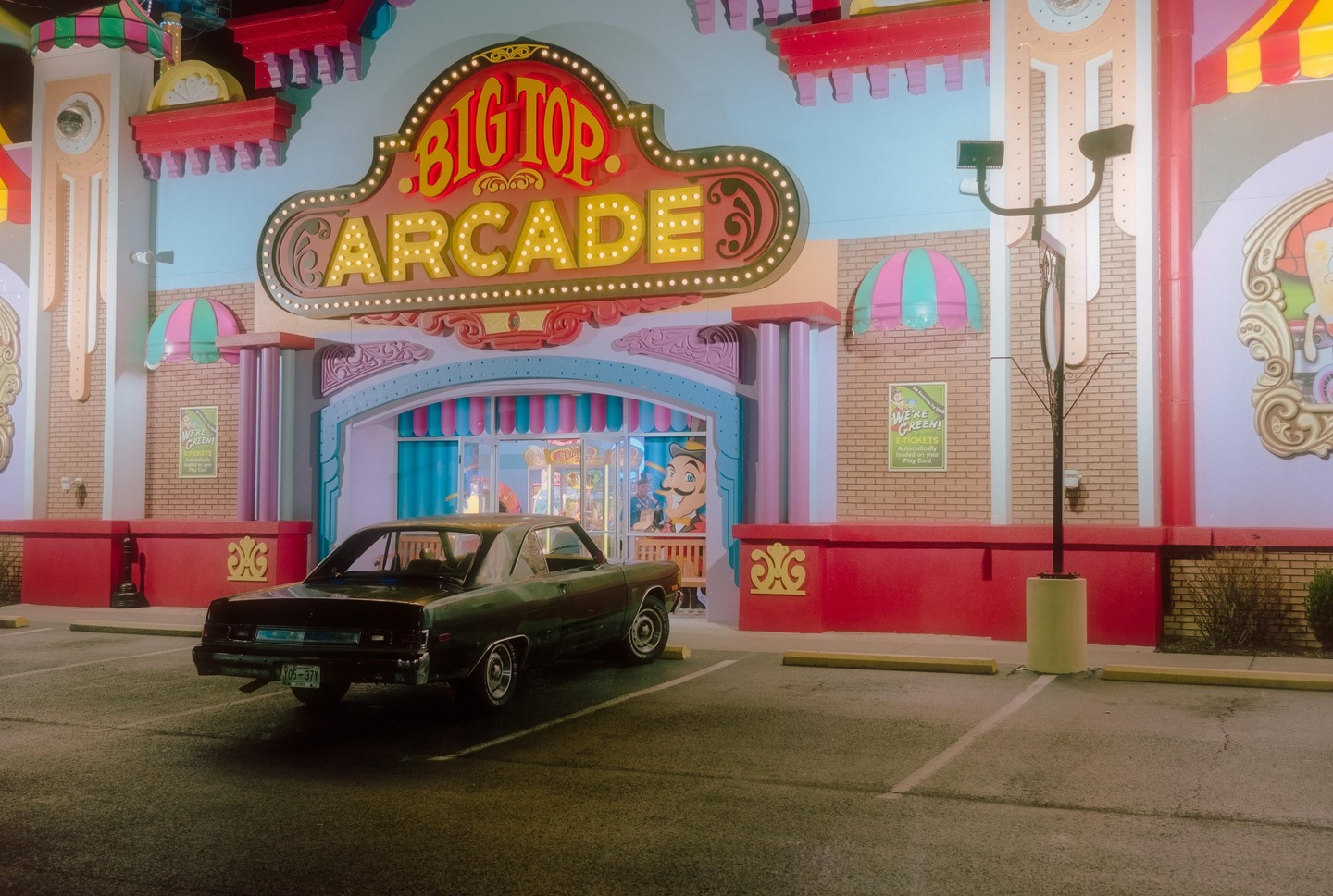 A dark green car is parked in front of the brightly lit "Big Top Arcade" building at night.