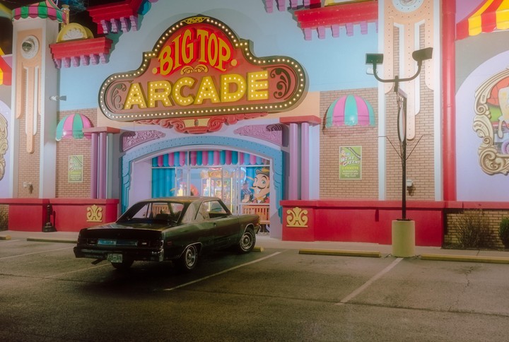 A dark green car is parked in front of the brightly lit "Big Top Arcade" building at night.