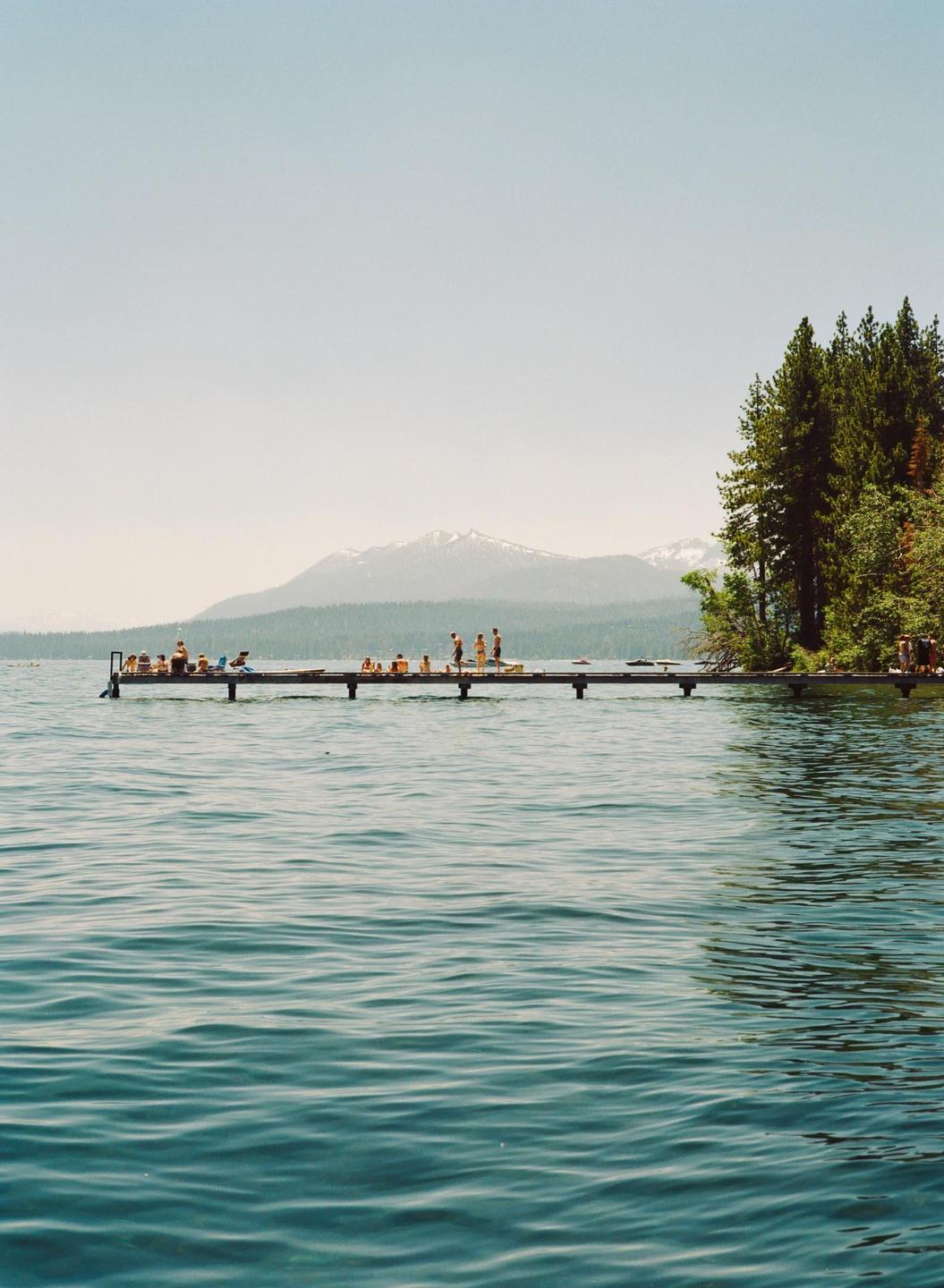 A long pier with many people on it extends into a lake with mountains and trees in the background.