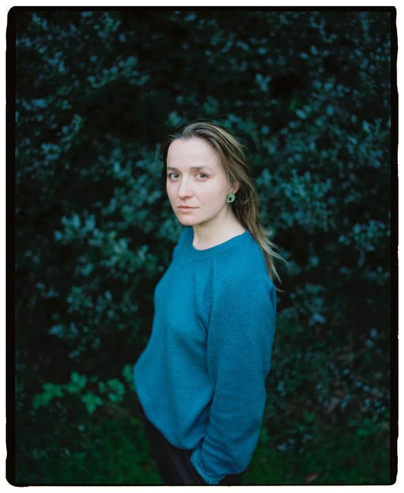 A woman with long hair and a blue sweater stands in front of a dark green bush.