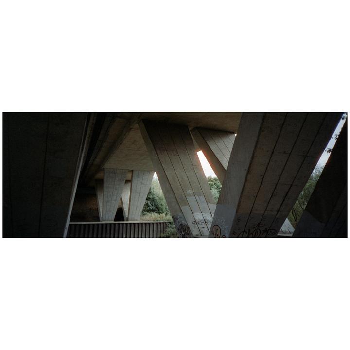 The concrete supports of a bridge are visible from below, with graffiti on some of the supports and trees in the background.