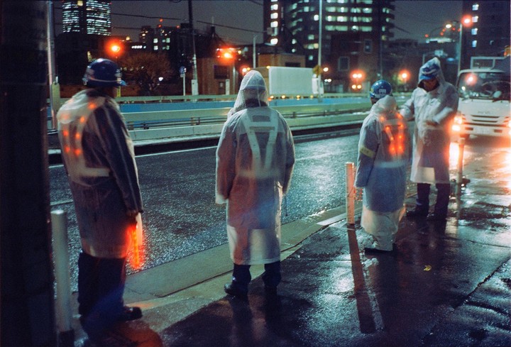 Three men in reflective raincoats stand on a wet street at night with city lights in the background.