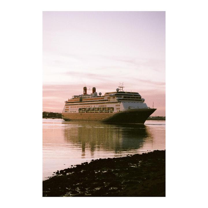 A large cruise ship with red smokestacks floats on calm water under a pink sky.