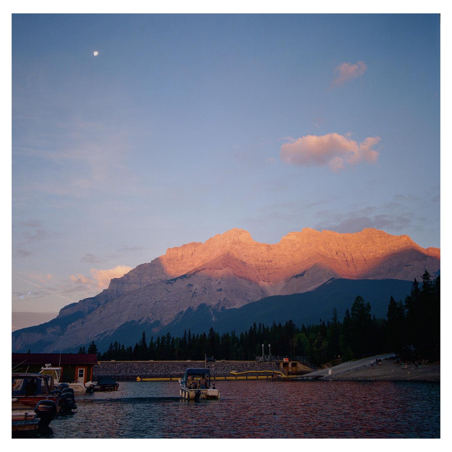 A moon is visible in the sky above a mountain with a lake and boats in the foreground.