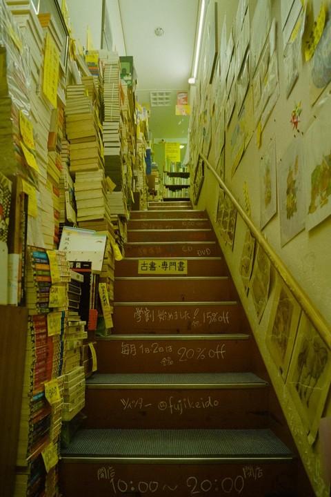 A narrow staircase in a bookstore is lined with tall stacks of books and posters on the walls, with Japanese writing on the stair risers.