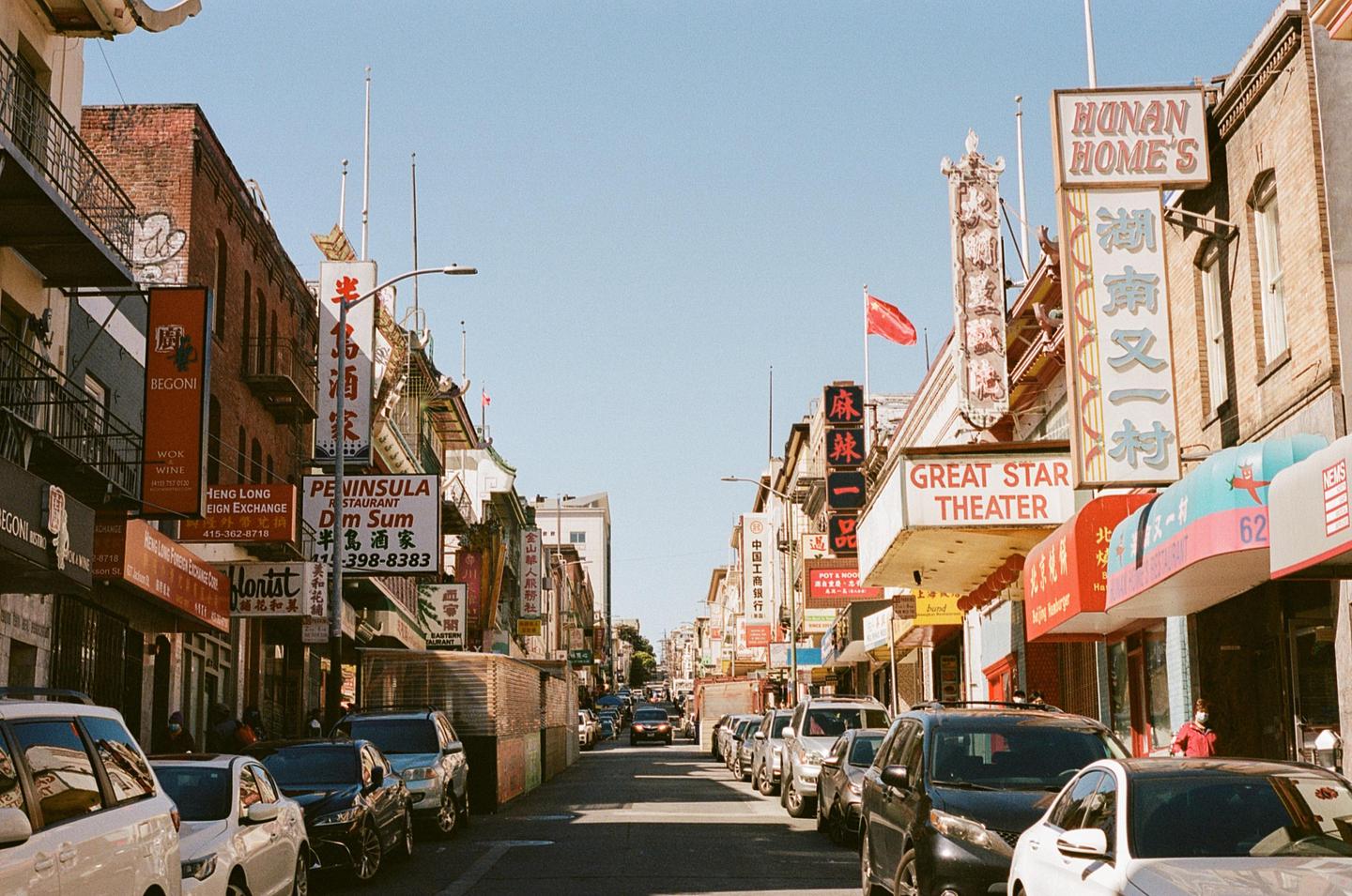 A street in Chinatown is lined with parked cars and buildings with many signs in English and Chinese.