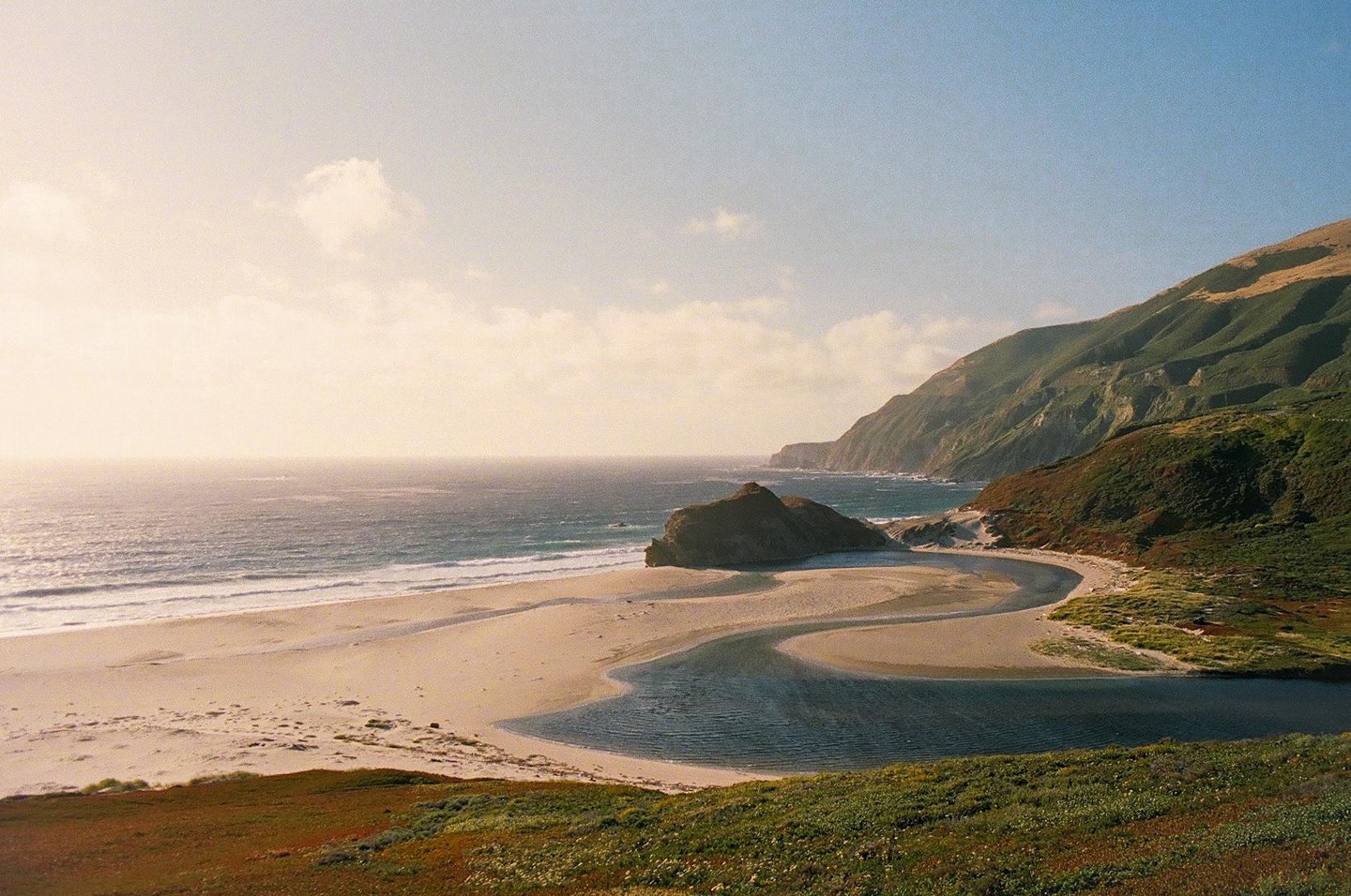 A winding river flows into the ocean next to a sandy beach and a large rock formation, with green hills rising in the background.