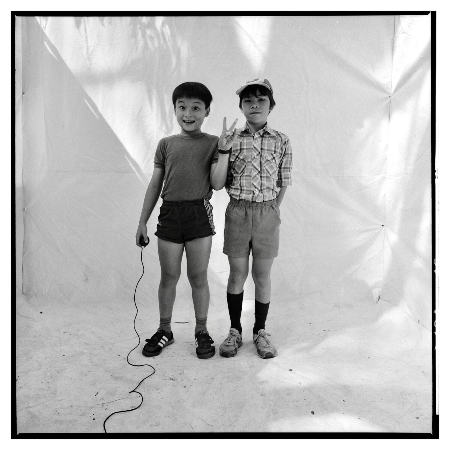 Two boys stand side by side in front of a white backdrop, one holding a jump rope and the other making a peace sign.