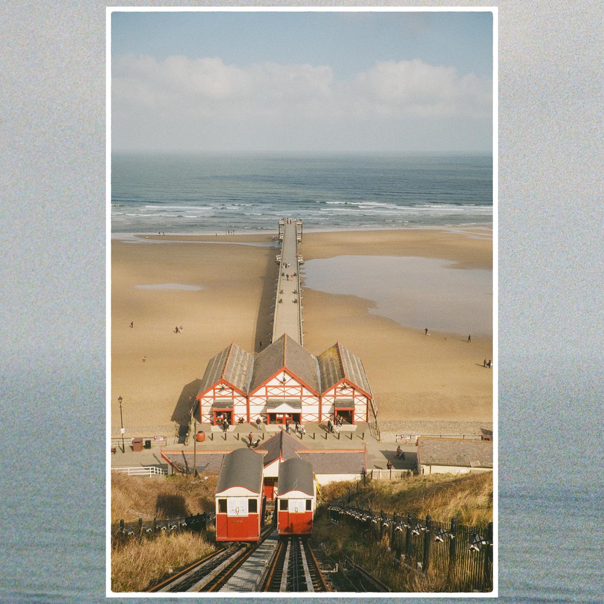 Two red funicular cars are on tracks leading down to a beach with a pier and buildings.