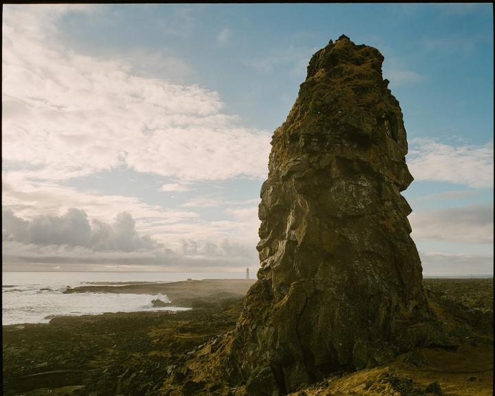 A large rock formation covered in moss stands prominently on a rocky coastline with a lighthouse in the distance under a cloudy sky.