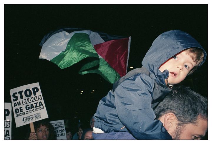 A child in a hooded jacket sits on a man's shoulders at night, with a Palestinian flag and protest signs visible in the background.