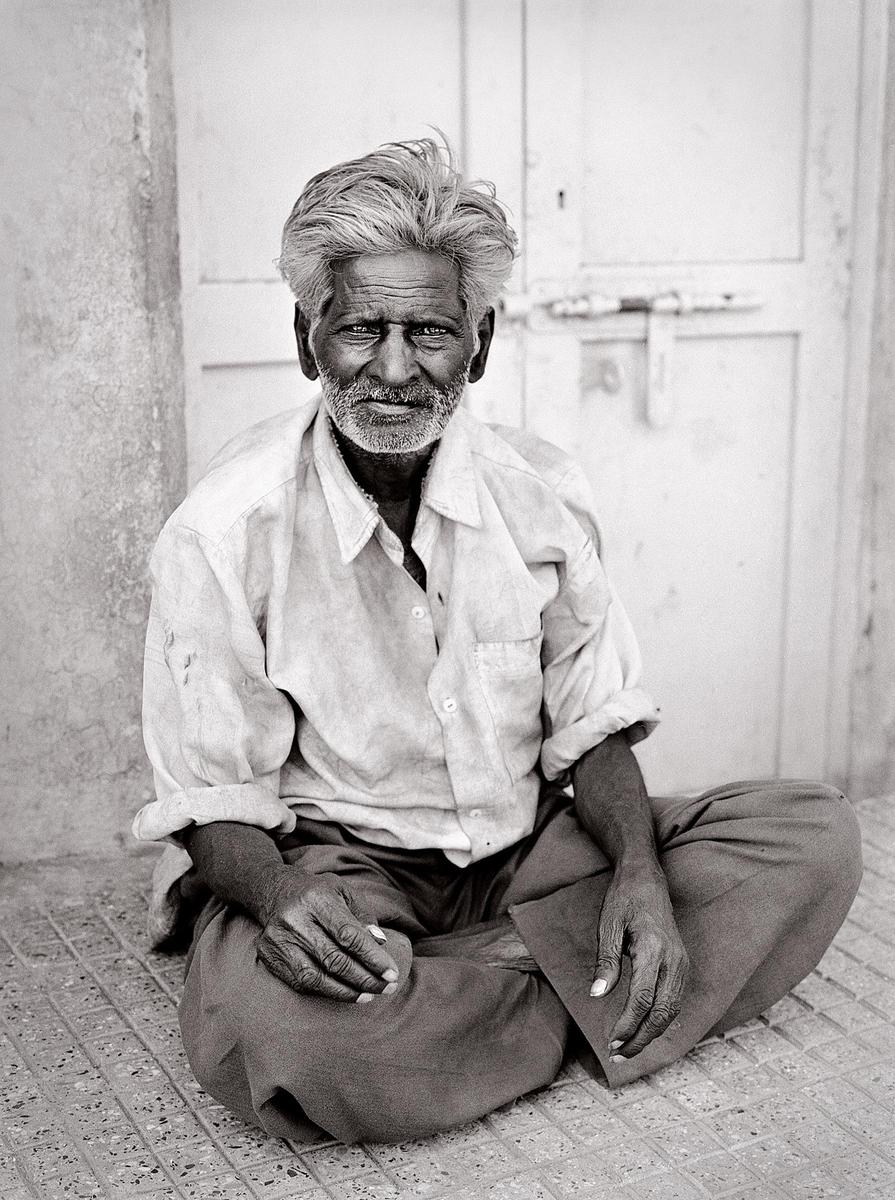 A man with gray hair and a beard sits cross-legged on a tiled surface in front of a door.