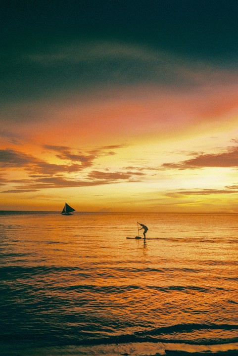 A person paddleboarding on the ocean at sunset with a sailboat in the distance.