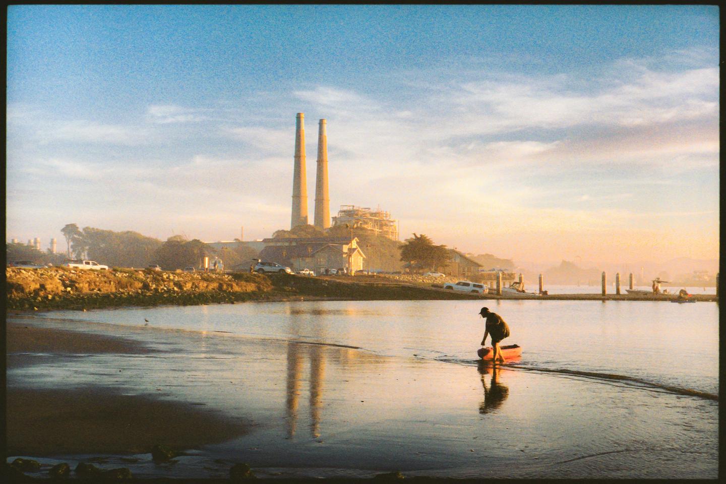 A person pushes a small boat into the water with a power plant and buildings on the shore in the background.