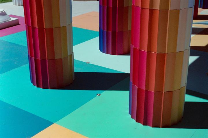 Three colorful pillars stand on a brightly painted ground with geometric patterns and shadows.