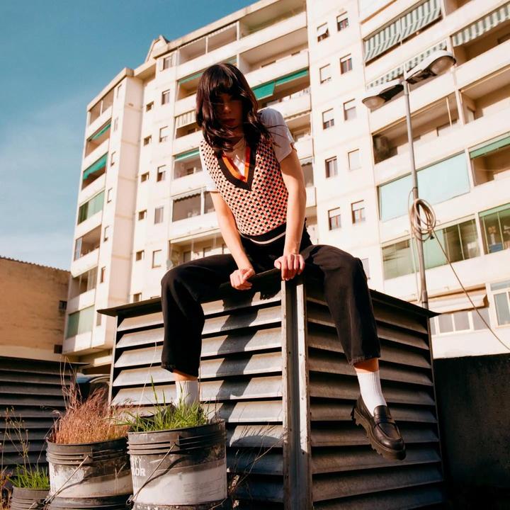 A woman sits on a metal structure on a rooftop with an apartment building in the background.